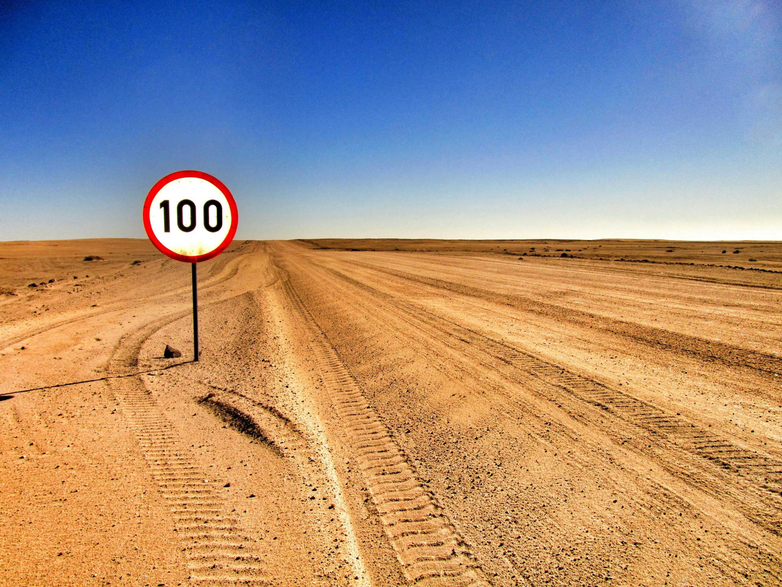 Open desert road with a speed limit sign showing 100 under clear sky.